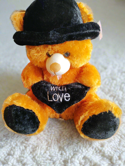 Teddy bear wearing a black hat and heart-shaped sign with 'With Love' text, in a store setting.